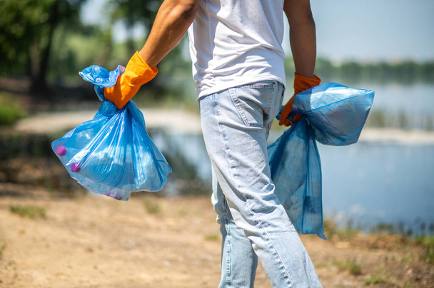 Garbage collection. Dark-skinned guy in gloves with back to camera holding bags of waste walking near river on sunny day - Zdjęcie, obraz