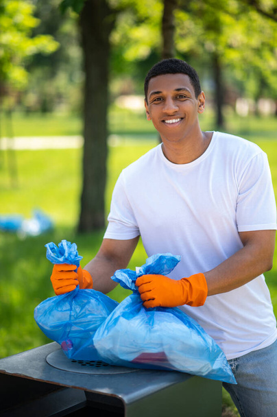 Lucky day. Dark-skinned guy in protective gloves smiling at camera holding waste bags over trash can in green park - 写真・画像