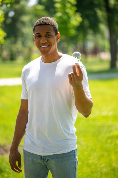 Protection of nature. Dark-skinned guy smiling at camera showing lightbulb standing in park on sunny day - Foto, Imagem