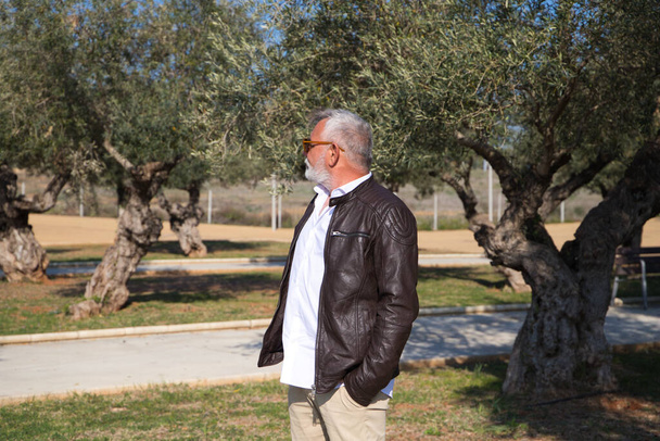 Przystojny, dojrzały mężczyzna z brodą i siwymi włosami spaceruje po parku. Mężczyzna jest ubrany w beżowe spodnie, białą koszulę i skórzaną kurtkę. Pojęcie wieku emerytalnego - Zdjęcie, obraz