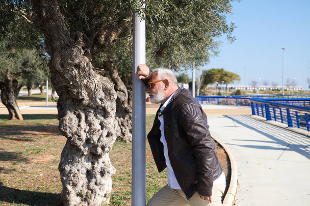 Handsome mature man with beard and grey hair is leaning against a lamppost pensively in the park. The man is dressed in beige trousers, white shirt and leather jacket. Old age concept - 写真・画像