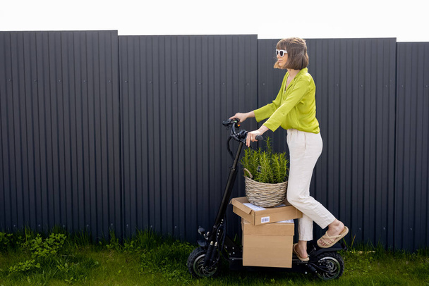 Joven mujer conduciendo scooter eléctrico con parcelas de cartón y planta verde sobre fondo de pared gris. Concepto de entrega y estilo de vida sostenible - Foto, imagen