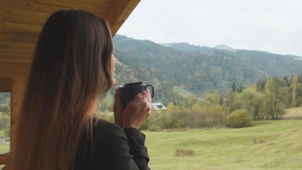 Long-haired beautiful woman holds cup and drinks coffee, tea, view mountains, forest, valley. Cloudy, medium shot. Concept of feedback, hotel, good morning, relaxation, enjoyment, nature. High quality - Filmati, video