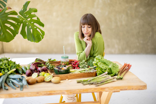 Eine junge Frau sitzt am Tisch voller frischer Lebensmittelzutaten in einem Raum mit grünen Pflanzen. Gesunde natürliche Ernährung und Lebensstil-Konzept - Foto, Bild