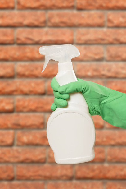 Woman's hand in a green protective glove with the bottle of glass cleaning liquid and a brick wall on the background. Washing and cleaning concept. - Foto, Bild