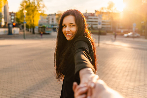 Happy brunetka dziewczyna z uśmiechem na twarzy trzymając chłopaka za rękę na ulicy o zachodzie słońca na ciepłe wiosnę, letni wieczór.Follow-me koncepcji.Skupienie. - Zdjęcie, obraz