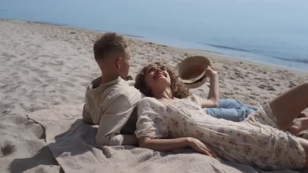 Carefree young couple lying sand beach summer time close up. Cheerful smiling girl with curly hair putting straw hat on man head. Lovely pair relax on beautiful seashore sunny day. Concept rest. - Video, Çekim