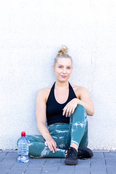 Sport and lifestyle concept, sporty blonde woman resting after doing sports outdoors. Attractive young blonde Caucasian woman relax after workout with happiness and joy, and a water bottle Full length - Foto, Imagem