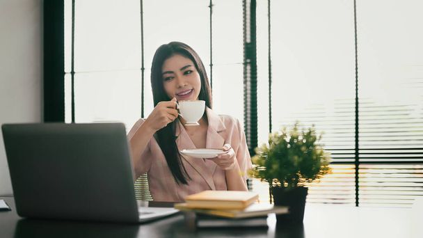 Agréable femme de travail buvant du café et lisant des nouvelles en ligne ou un courriel d'affaires sur son ordinateur portable. - Photo, image