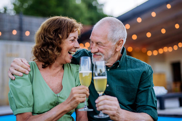 A man with number 70 balloons with his wife celebrating birthday and toasting with wine near backyard pool. - 写真・画像