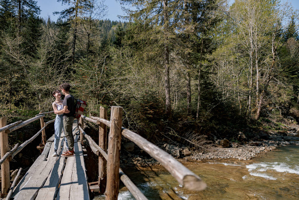 Couple in love standing on wooden bridge and hugging over fast river. Rural nature. Trekking. - Фото, изображение