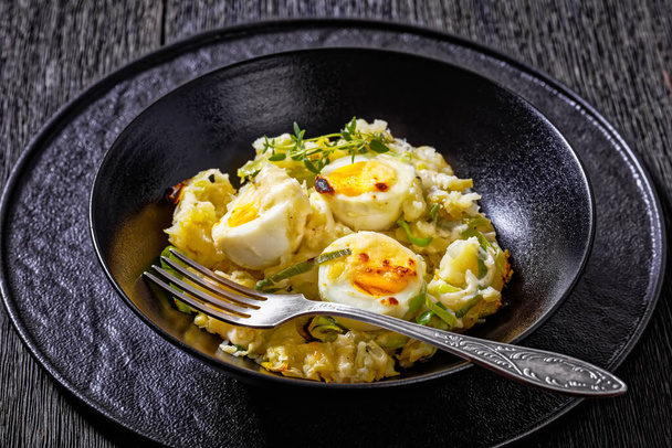 Welsh Anglesey Eggs, soft mashed potatoes with hard-boiled eggs all smothered in thick creamy leek and cheese sauce in black bowl on dark oak table, horizontal view, close-up - Fotoğraf, Görsel