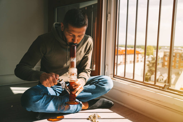 Man smoking bong and inhaling the smoke at home. Man smoking pot, medical marijuana or cannabis from a bong or water pipe. Cannabis and weed legalisation concept. High quality photo - Foto, immagini