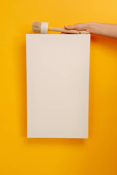 Hand holding white canvas with copy space and wooden paint brush tool on yellow background. Empty sheet banner with place for text. Concept of design, art creativity - Φωτογραφία, εικόνα