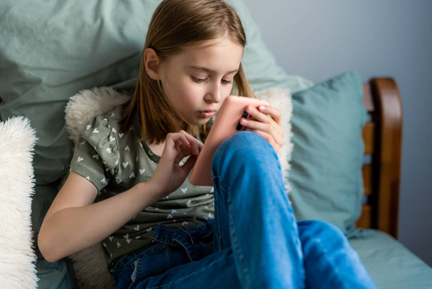 Pretty preteen girl sitting in the bed and looking at smartphone at home in the bedroom. Beautiful kid with cell phone indoors - Fotografie, Obrázek