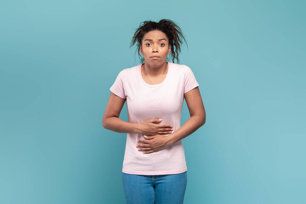 Ból brzucha, wzdęcia i mdłości. Młoda czarna dama dotykająca bolącego żołądka po jedzeniu, stojąca nad niebieskim tłem studia. Ból brzucha, zapalenie brzucha - Zdjęcie, obraz