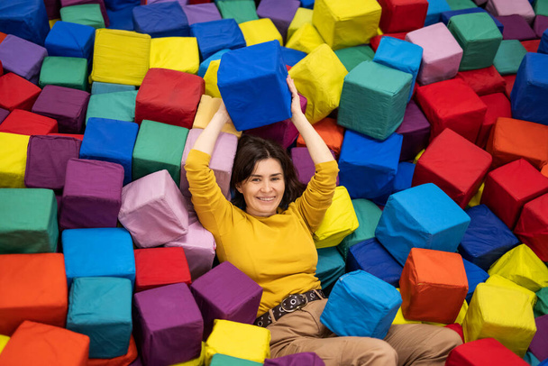 Pretty girl lying on trampoline and holding colorful cube. Young woman enjoying entertaiments at playground park - Foto, afbeelding