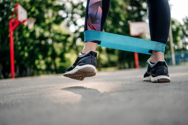 Young girl woman exercising outdoors with mini rubber elastic band doing workout. Closeup view of female person legs during active training with additional sport equipment outside - Valokuva, kuva