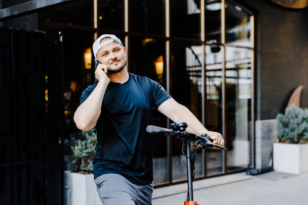 Caucasian young handsome man standing on street at electric scooter and talking on mobile phone. - Фото, изображение