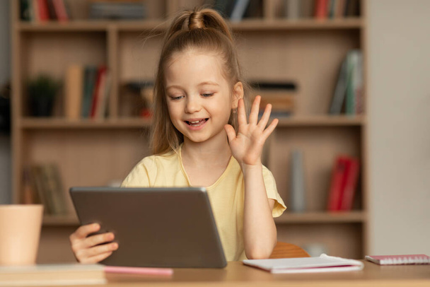 Schoolmeisje zwaaien Hallo aan digitale tablet maken van video bellen en leren Online zitten aan de balie thuis. Meisje communiceert met behulp van computer. Externe communicatie en onderwijs Concept - Foto, afbeelding