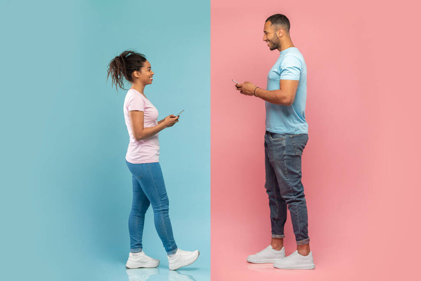 Positive african american spouses holding smartphones and looking at each other, using cellphones while walking over blue and pink studio background, side view - Фото, зображення