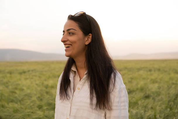 Smiling woman, beautiful long haired brunette smiling woman. Standing in the green wheat field. Attractive caucasian millennial female portrait at sunset. Modern woman farmer or agronomist concept. - Fotó, kép