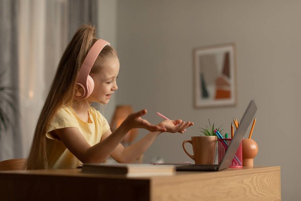 Маленькая девочка делает видеозвонок на ноутбуке Разговаривая с компьютером веб-камера обучения в Интернете, сидя за столом дома. Ребенок, имеющий дистанционное общение класса с помощью гаджетов. Шот с видом сбоку - Фото, изображение