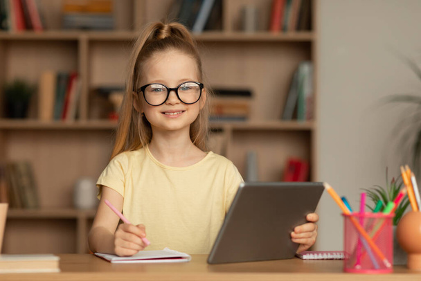 Schoolgirl Taking Notes Using Digital Tablet Posing Smiling To Camera Sitting At Desk Indoors. Kid Learning Online Doing Homework At Home. Remote Internet Education Concept - 写真・画像