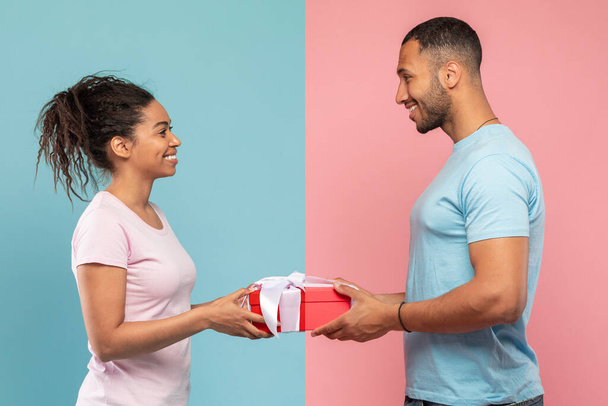 Loving black man giving gift to his wife, posing over blue and pink studio background, side view. African americn guy giving present to his girlfriend. Valentines day, anniversary celebration - 写真・画像