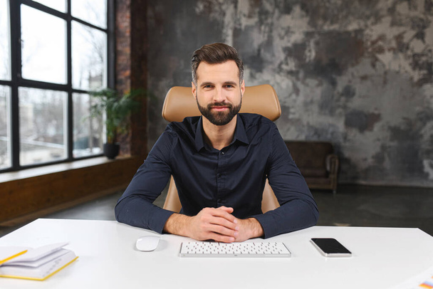Portrait of handsome young businessman in smart casual wear sitting at the desk in loft office and looks at camera, male employee involved virtual meeting, webcam view - Zdjęcie, obraz