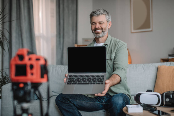 Happy friendly senior european man blogger with beard records video review on technology, shows laptop with empty screen in room interior. Modern work, hobby and vlog on devices at home in free time - 写真・画像