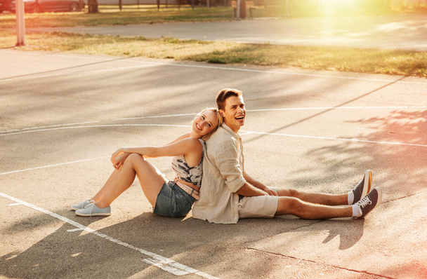summer holidays and people concept - happy young couple sitting back to back on basketball playground - Zdjęcie, obraz