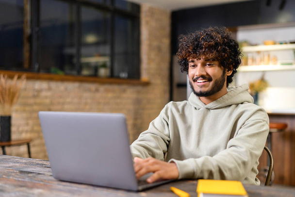 Happy smiling man distantly working from home. Successful male freelancer sitting at the desk, using laptop, develop a project. Indian student on a lecture. Online study concept - 写真・画像