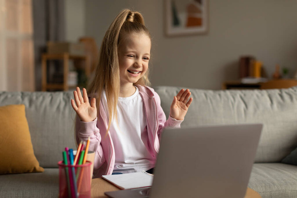 Електронне навчання. Весела школярка робить відео виклик Wave Hello To Laptop Computer Learning Online, сидячи на дивані вдома. Інтернет школа освіта та послуги концепція
 - Фото, зображення