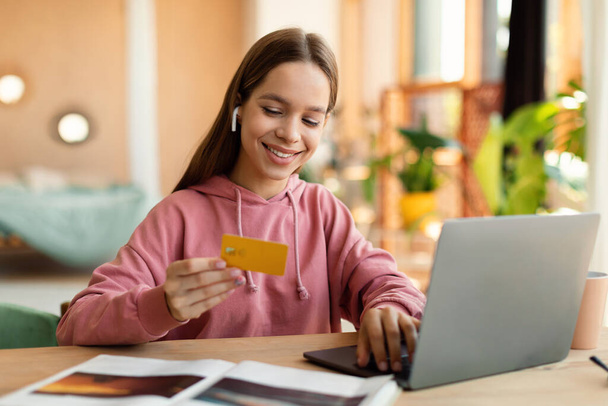 Happy teenage girl using laptop and credit card, sitting at desk and ordering goods online from home, copy space. Positive adolescent buying things on web - Foto, imagen