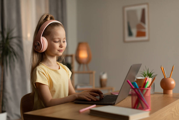 Schulmädchen, die am Laptop online lernen und Kopfhörer tragen, sitzen zu Hause am Schreibtisch. Kleines Mädchen beim Hausaufgabenmachen im Internet auf dem Computer. Technologie und Bildung. Seitenansicht - Foto, Bild
