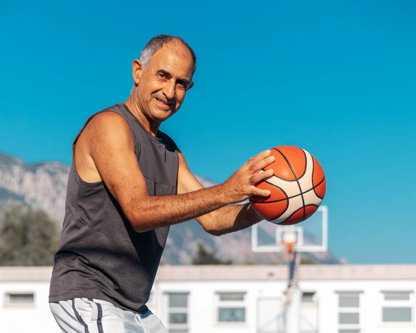 Half-length portrait of 60-years old smiling basketball player in good shape ready to shoot or pass a sport ball outdoor - Photo, image