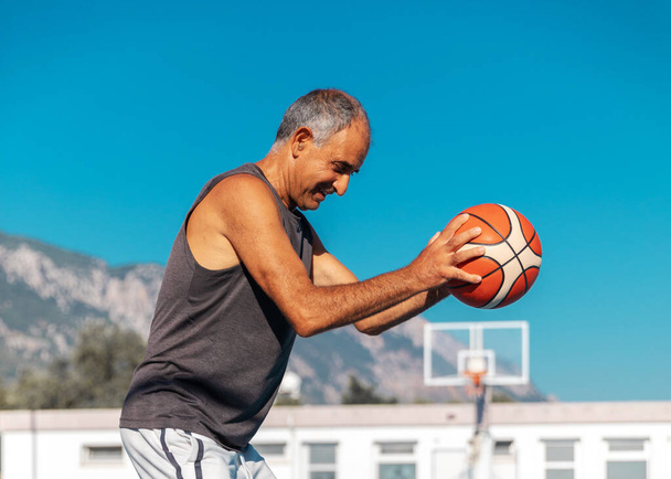60 yaşındaki gülümseyen basketbolcunun yarı boy portresi. Açık havada elinde spor topu tutuyor. - Fotoğraf, Görsel