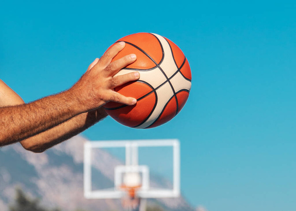 Yetişkin bir adamın elleri bir basketbol topunu tutuyor ve güneşli yaz günlerinde mavi gökyüzünde pas vermeye hazır.. - Fotoğraf, Görsel