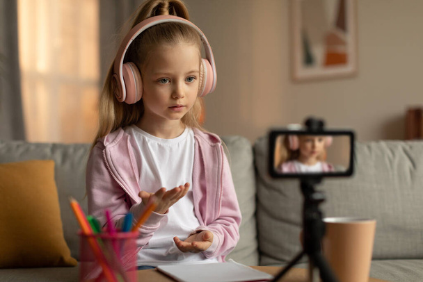 E-Learning. Little Schoolgirl Learning Online Video Calling Via Cellphone On Tripod Having Remote Class At Home. Girl Wearing Headphones Watching Video Lesson Indoor. Selective Focus - Φωτογραφία, εικόνα