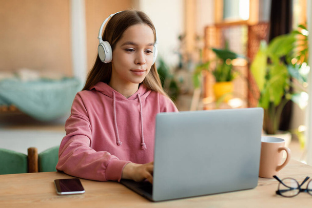 Positive schoolgirl in headphones watching video lesson remotely on laptop, sitting at desk at home interior. Schooling, modern education and knowledge in internet - Foto, afbeelding