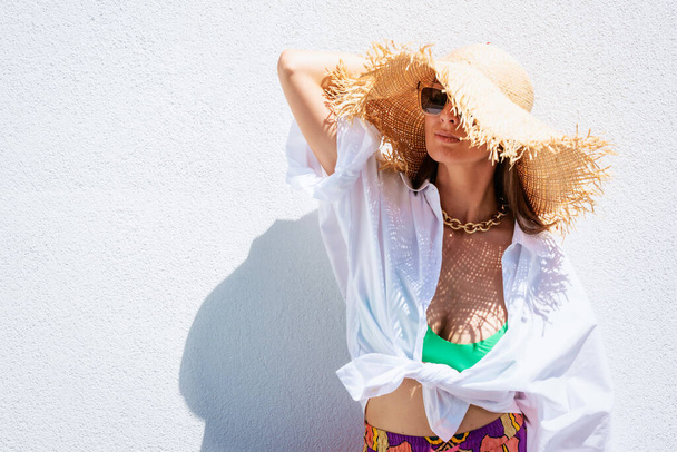 Portrait shot of an attractive woman wearing straw hat and sunglasses while standing at white wall. Happy woman enjoying sunshine. - Foto, imagen