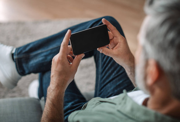 Elderly european man with beard plays in online game or watches video on smartphone with blank screen in room interior. Device for pensioner at home, new app and chat in social networks, ad and offer - 写真・画像