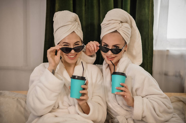 Két fiatal nő fürdőköpenyben és fürdőköpenyben, kávéscsészékkel a kanapén reggel.. - Fotó, kép