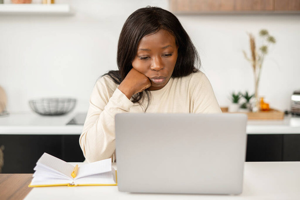 Sad young unemployed african american woman using laptop computer for job searching, sitting in home office and resting chin on hand, feels despair and depression - Photo, image