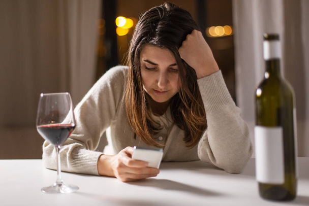 alcoholism, alcohol addiction and people concept - drunk woman or female alcoholic with smartphone drinking red wine at home - Фото, изображение