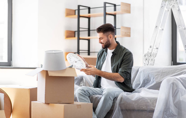 moving, people and real estate concept - happy smiling man with boxes and clock at new home - Fotoğraf, Görsel