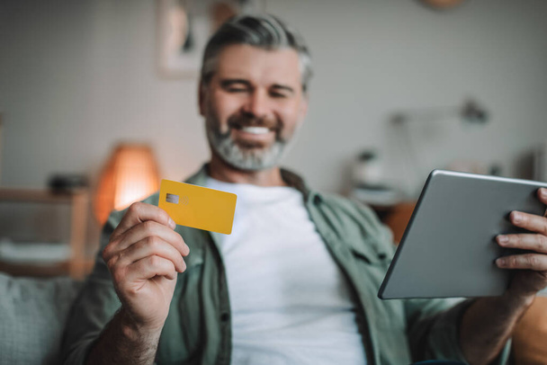 Happy senior european man with beard shopping online, holding tablet and credit card, enjoy huge sale in room interior. Banking and finance, money and app for shopaholic, gadget and social distance - Foto, Imagem
