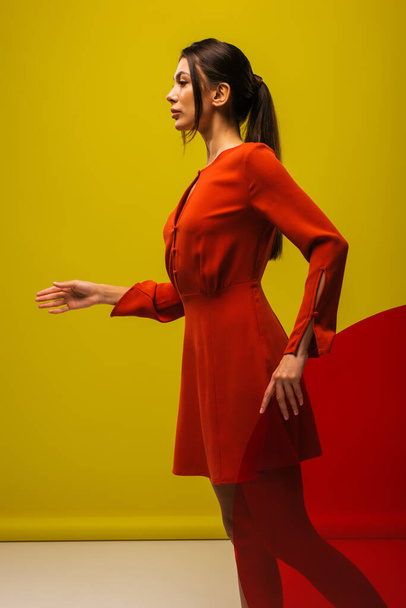 side view of stylish young woman in red dress holding round shape glass on green  - Foto, imagen