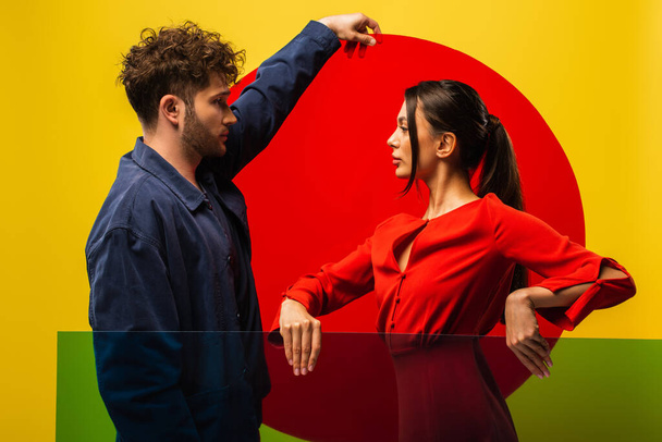 side view of trendy couple holding different shapes glass while posing isolated on yellow - Photo, image